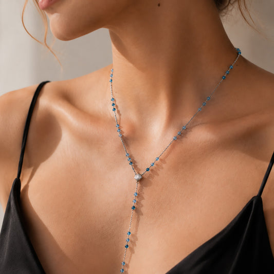 Close-up of a delicate white gold-tone Y-necklace featuring a seashell motif and a cascading drop of gradient blue gemstones.