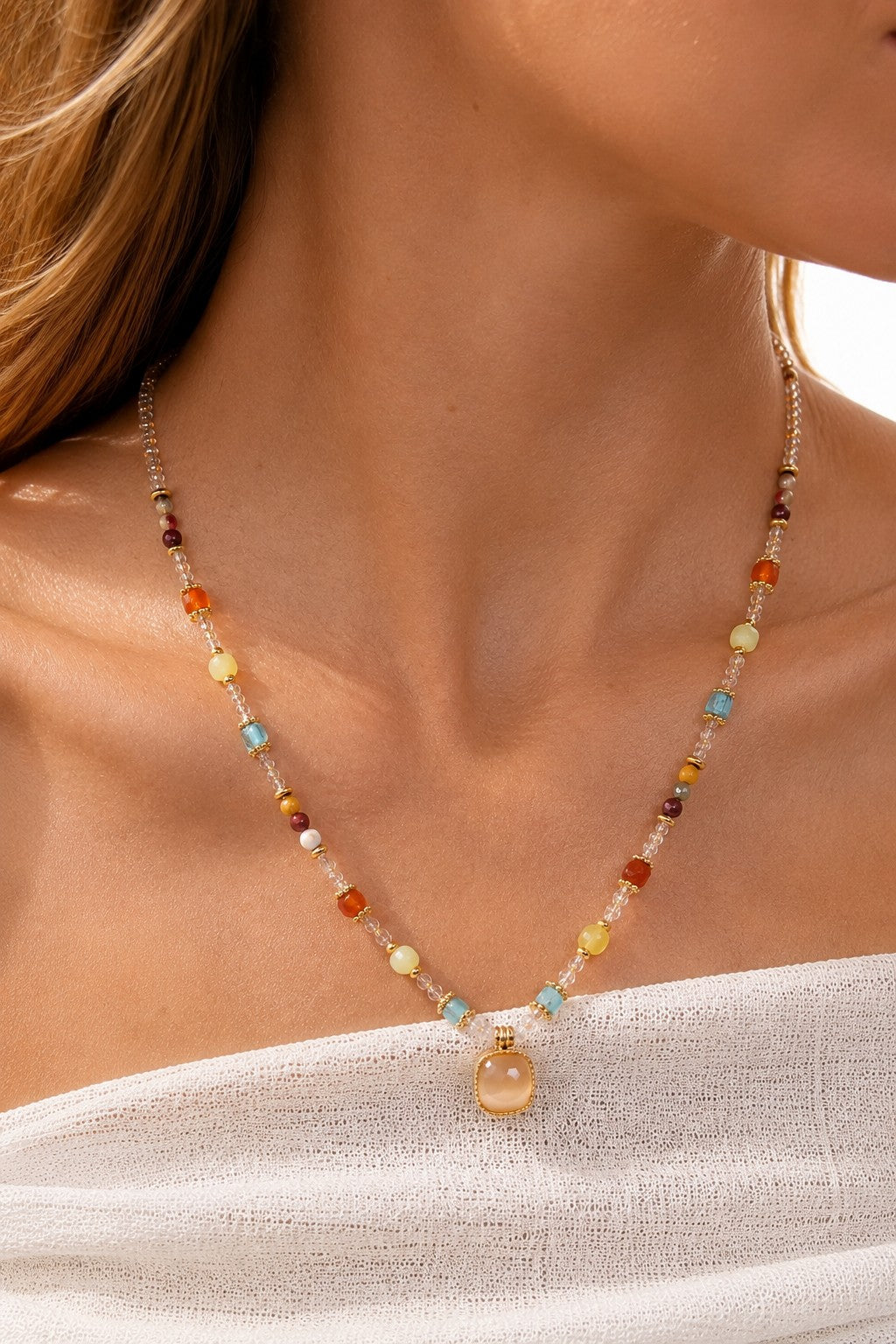 Close-up of a blonde model wearing a sun-drenched beaded gemstone necklace with an 18K gold cushion pendant against a white top.