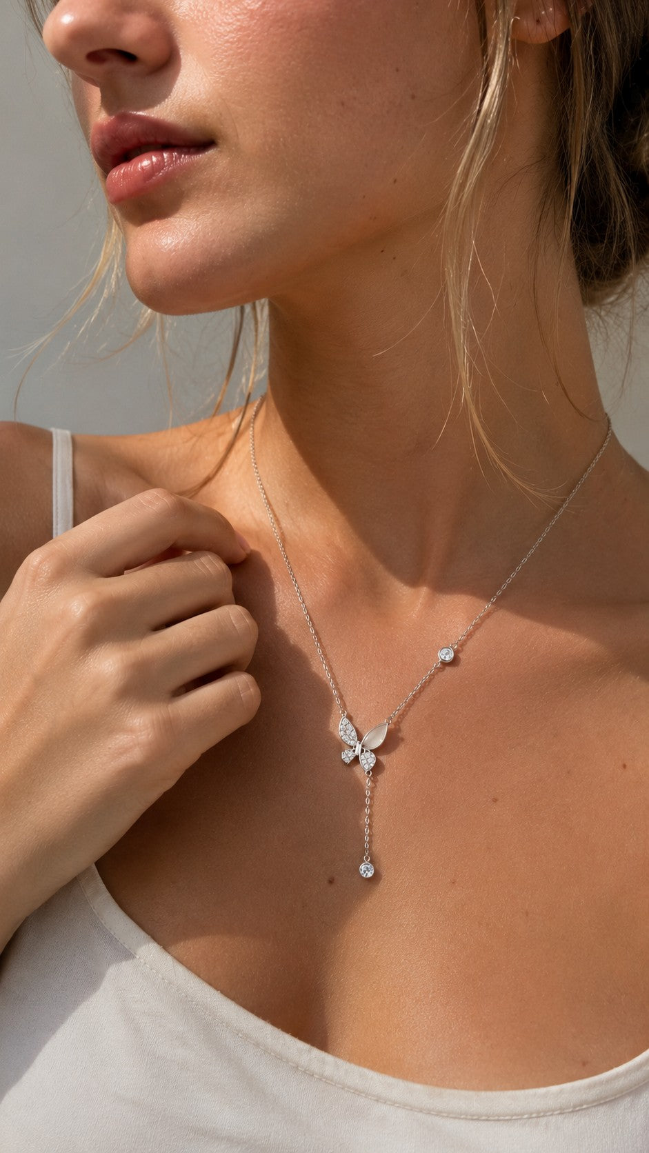 Close-up of a blonde model wearing a silver butterfly lariat Y-necklace with a white V-neck top under warm sunlight.