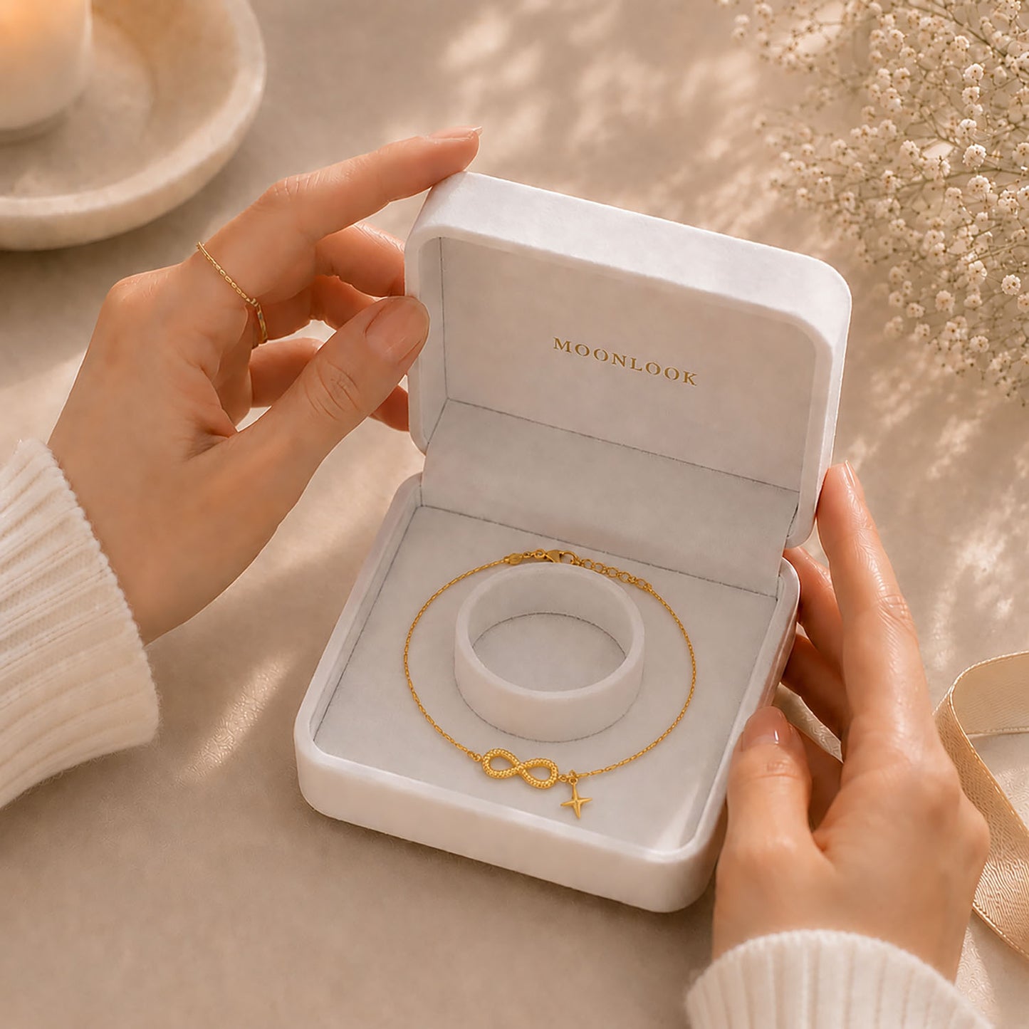 Hands gently opening a white Moonlook gift box containing a delicate gold-tone infinity and star anklet.
