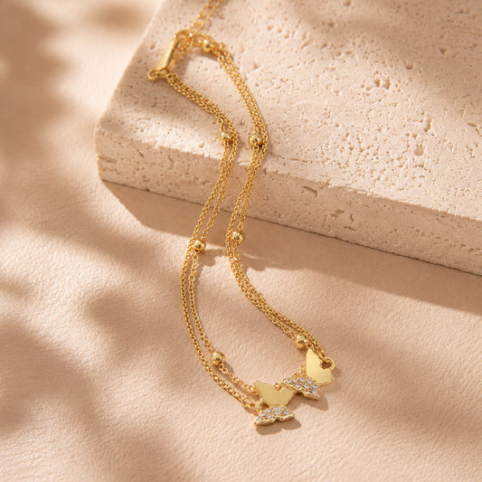 Elegant gold-tone double butterfly anklet draped over a textured stone block in warm sunlight.