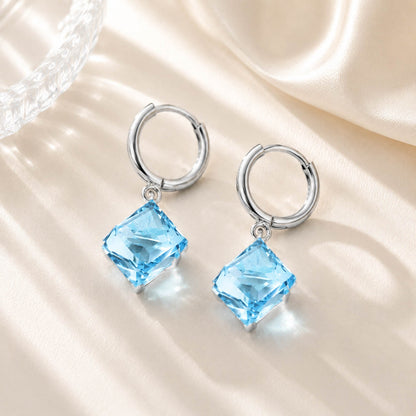 Close up of sparkling ocean blue square zirconia earrings resting on a soft silk and glass background in warm light.