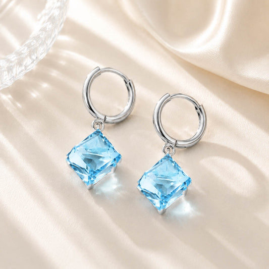 Close up of sparkling ocean blue square zirconia earrings resting on a soft silk and glass background in warm light.