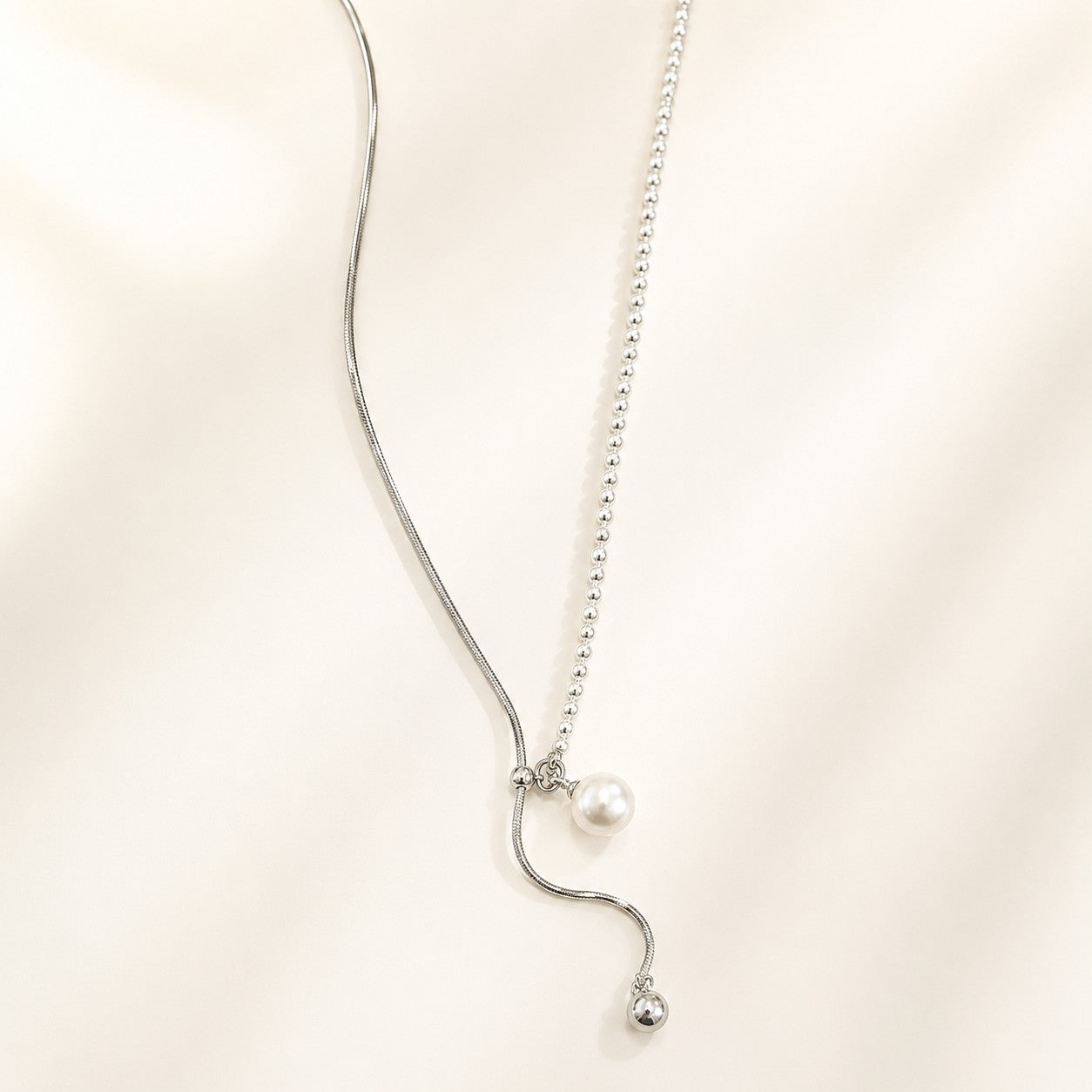 White gold plated asymmetric lariat necklace featuring a shell pearl and sliding metal ball drop chain laid on cream background