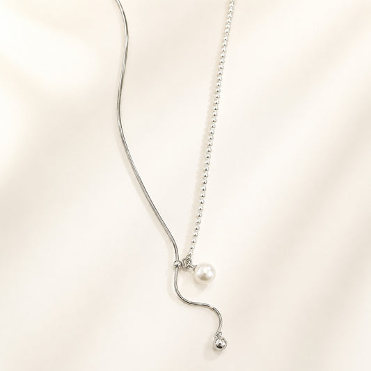 White gold plated asymmetric lariat necklace featuring a shell pearl and sliding metal ball drop chain laid on cream background