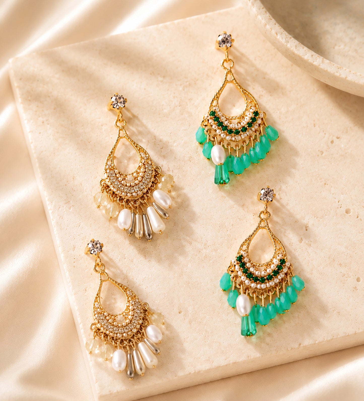 A beautiful display of gold-tone bohemian teardrop fringe earrings in both emerald green and pearl white colors on a textured stone surface.