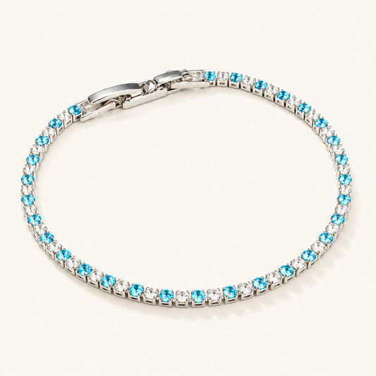 Minimalist flat lay of a white gold-tone tennis bracelet featuring alternating blue and clear zirconia on a clean white background.