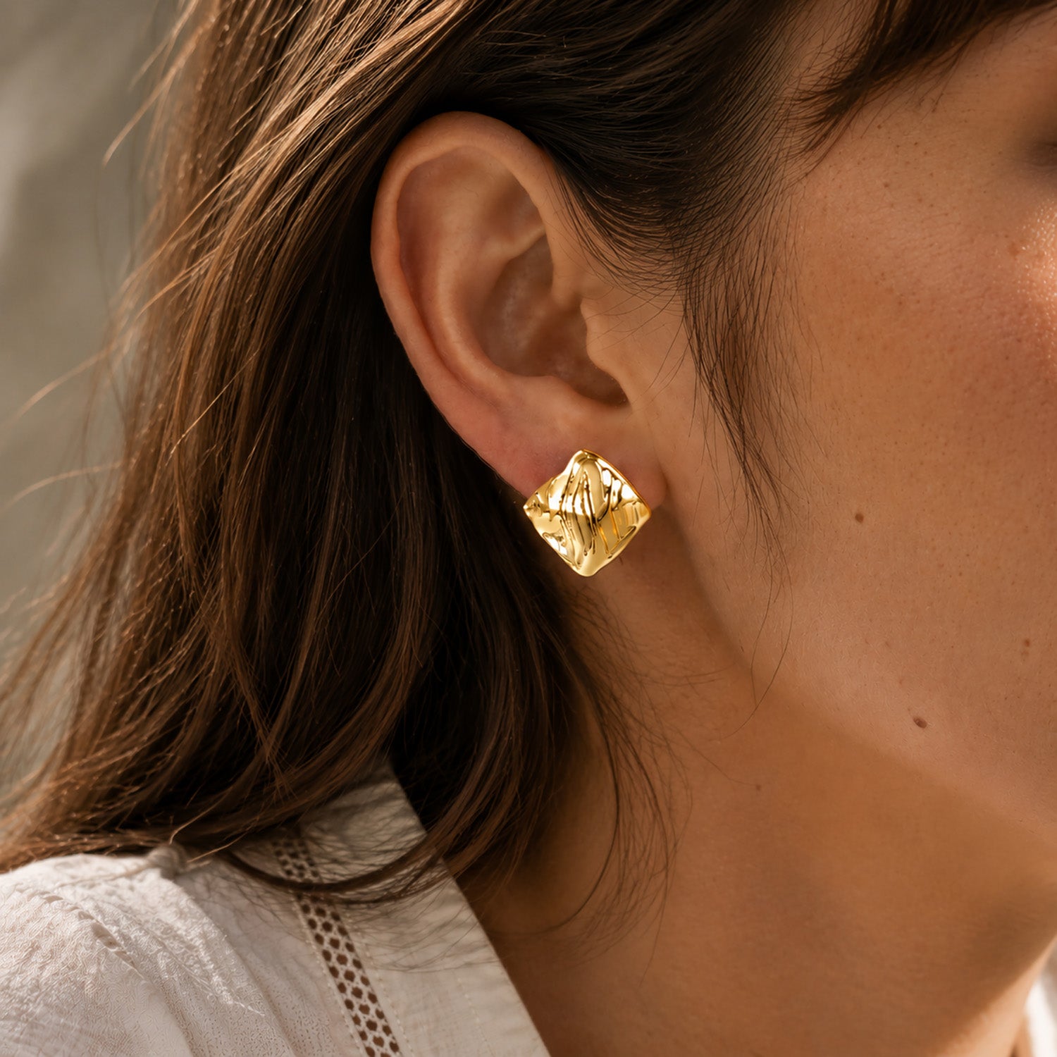 Macro close-up of a modern woman styling chunky geometric square stud earrings with a unique folded gold texture.