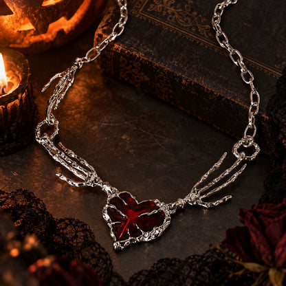 Flat lay of skeleton hand crimson heart pendant resting on antique book beside dark red roses