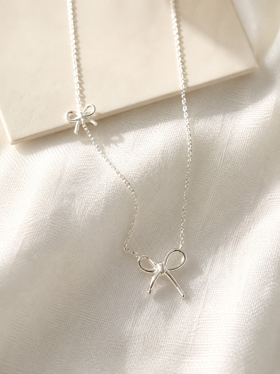 S925 sterling silver ribbon bow necklace resting on a soft white linen fabric under warm natural light.