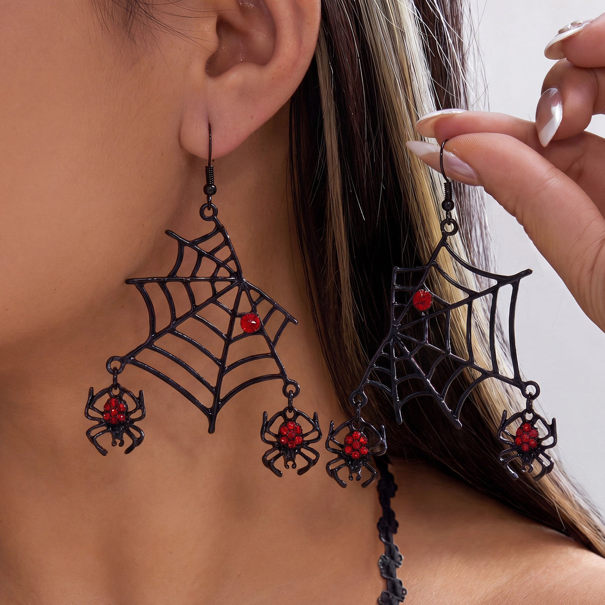 Close-up of model holding black spiderweb dangle earrings with hanging red rhinestone micro spiders