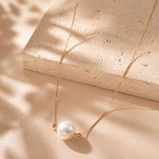 Elegant single pearl pendant necklace with a gold-tone chain laid on textured stone and silk.