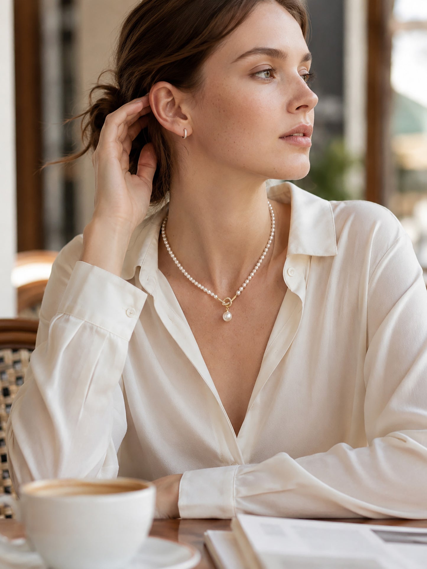Elegant woman in a crisp white shirt wearing a knotted pearl necklace at a cafe.