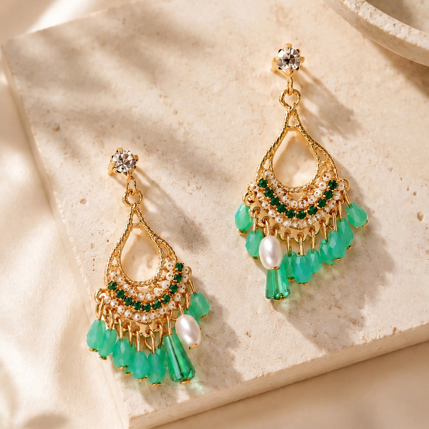 Close up of intricate gold-tone bohemian dangle earrings with green crystals resting on a warm stone surface.