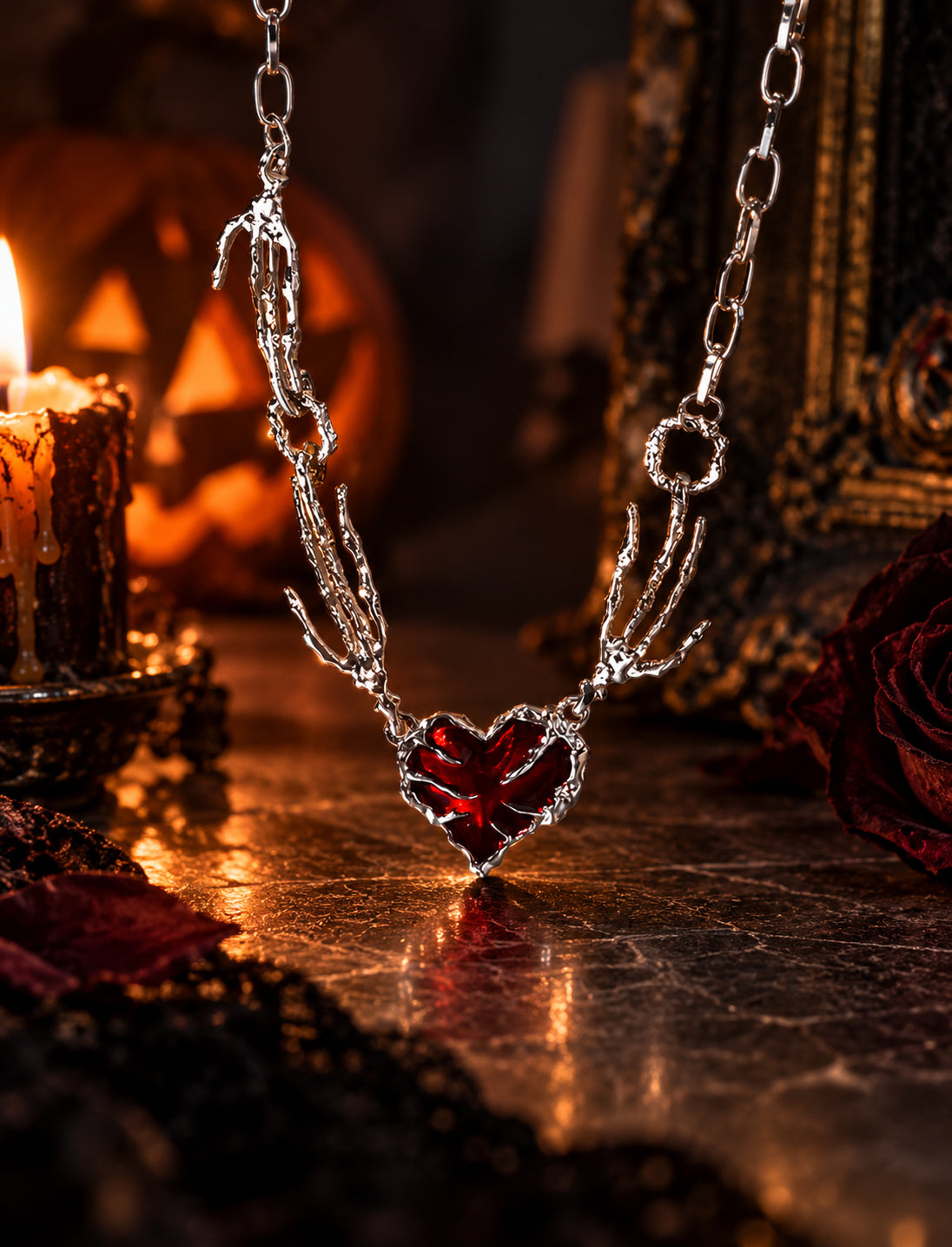 Close-up of gothic skeleton hand holding crimson red heart pendant necklace in front of lit Halloween pumpkin