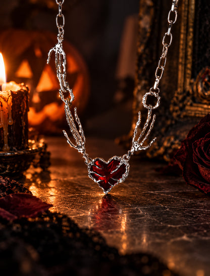 Close-up of gothic skeleton hand holding crimson red heart pendant necklace in front of lit Halloween pumpkin