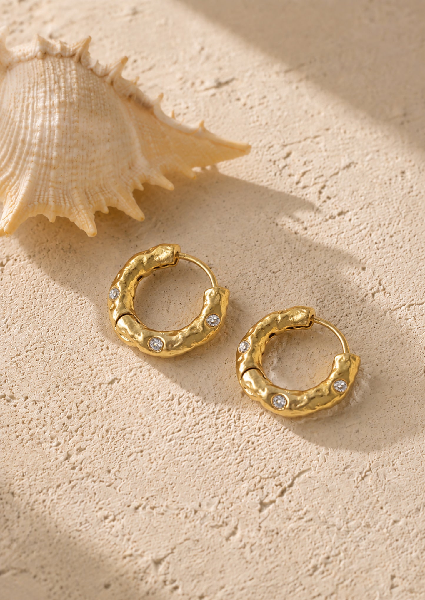 Artistic flat lay of textured gold-tone hoop earrings next to a natural seashell, highlighting their organic, coastal-inspired aesthetic.