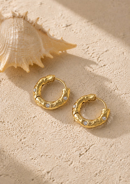 Artistic flat lay of textured gold-tone hoop earrings next to a natural seashell, highlighting their organic, coastal-inspired aesthetic.