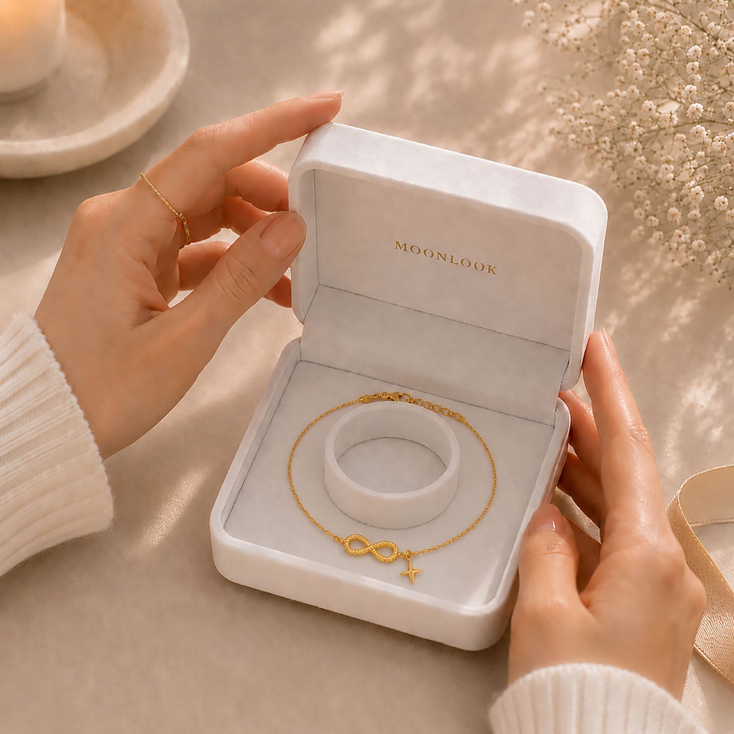 Hands gently opening a white Moonlook gift box containing a delicate gold-tone infinity and star anklet.