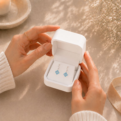 Hands opening a premium white Moonlook jewelry box revealing delicate ocean blue zirconia drop earrings.