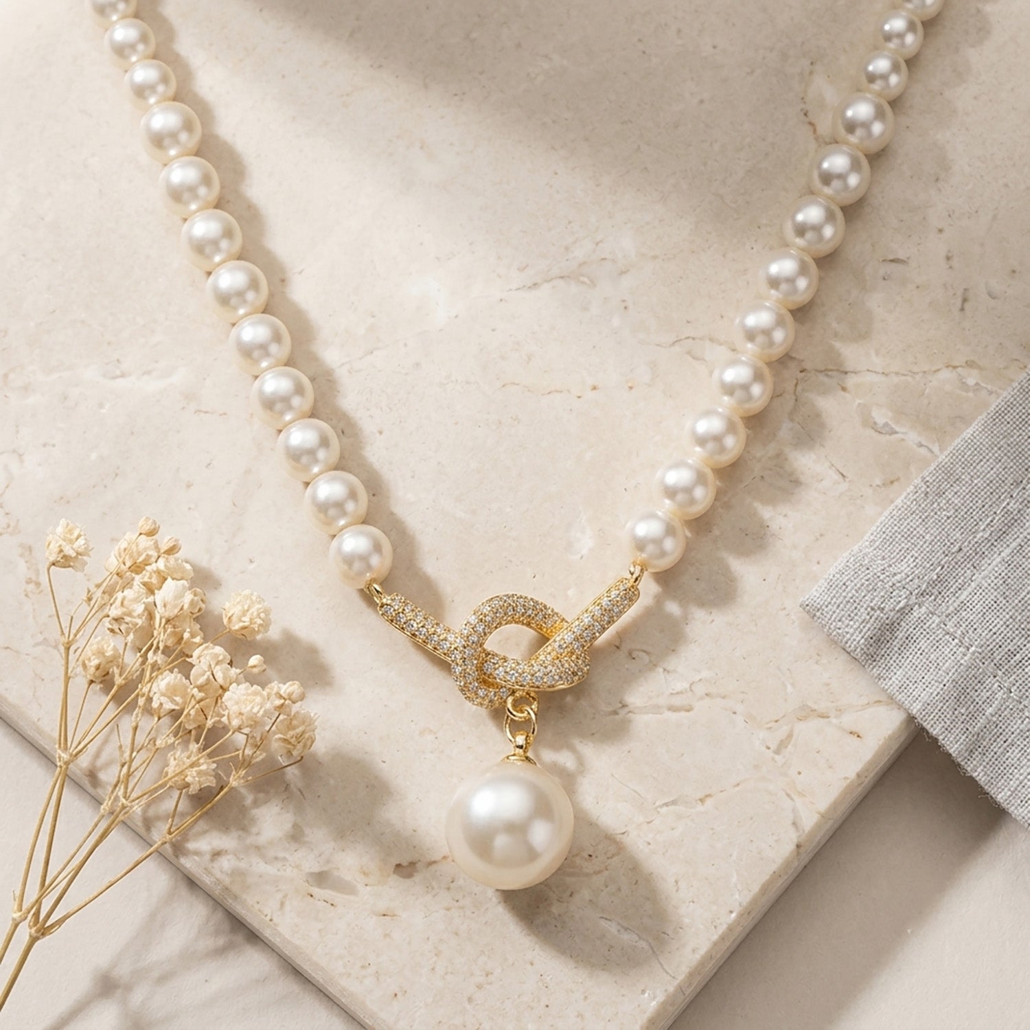 18K gold-tone knotted pearl pendant necklace laid flat on a stone surface with dried flowers.