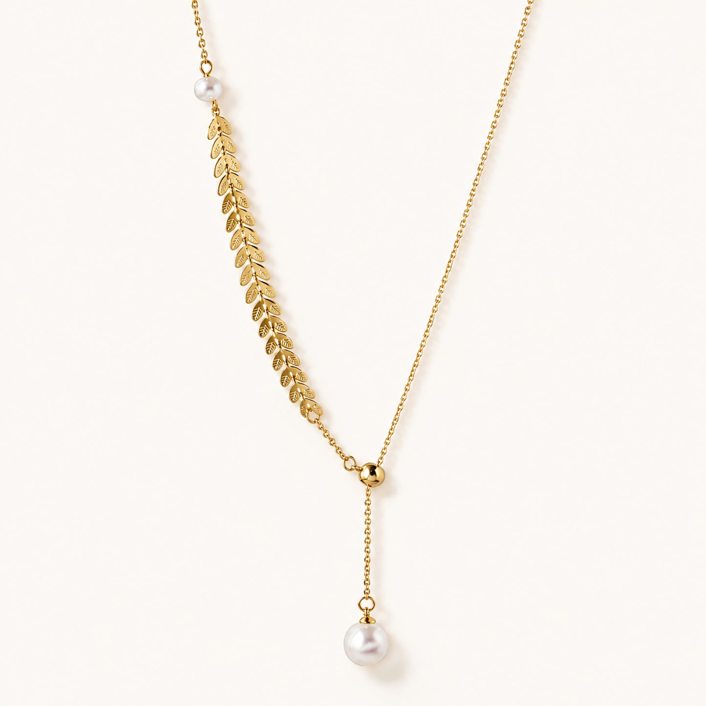 Minimalist flat lay of an elegant 18K gold-tone lariat necklace featuring a detailed leaf motif and pearl accents on a white background.