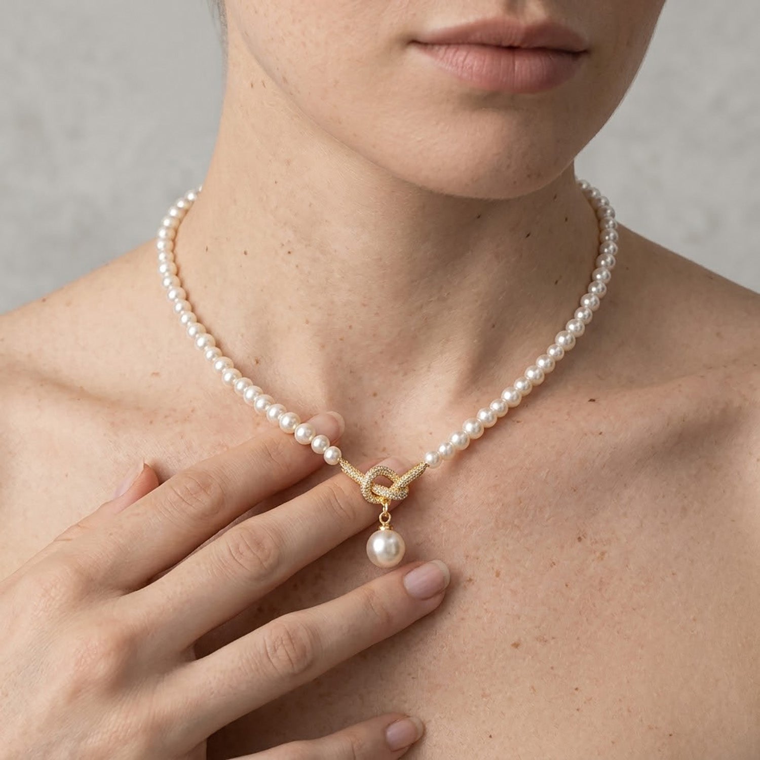 Close up of a woman touching an elegant 18K gold-tone knotted pearl pendant necklace on her collarbone.