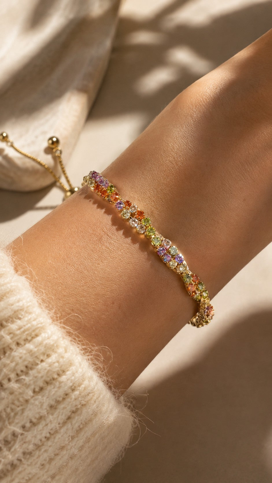 Close up of model wrist wearing adjustable twisted gold rainbow cubic zirconia tennis bracelet with sweater