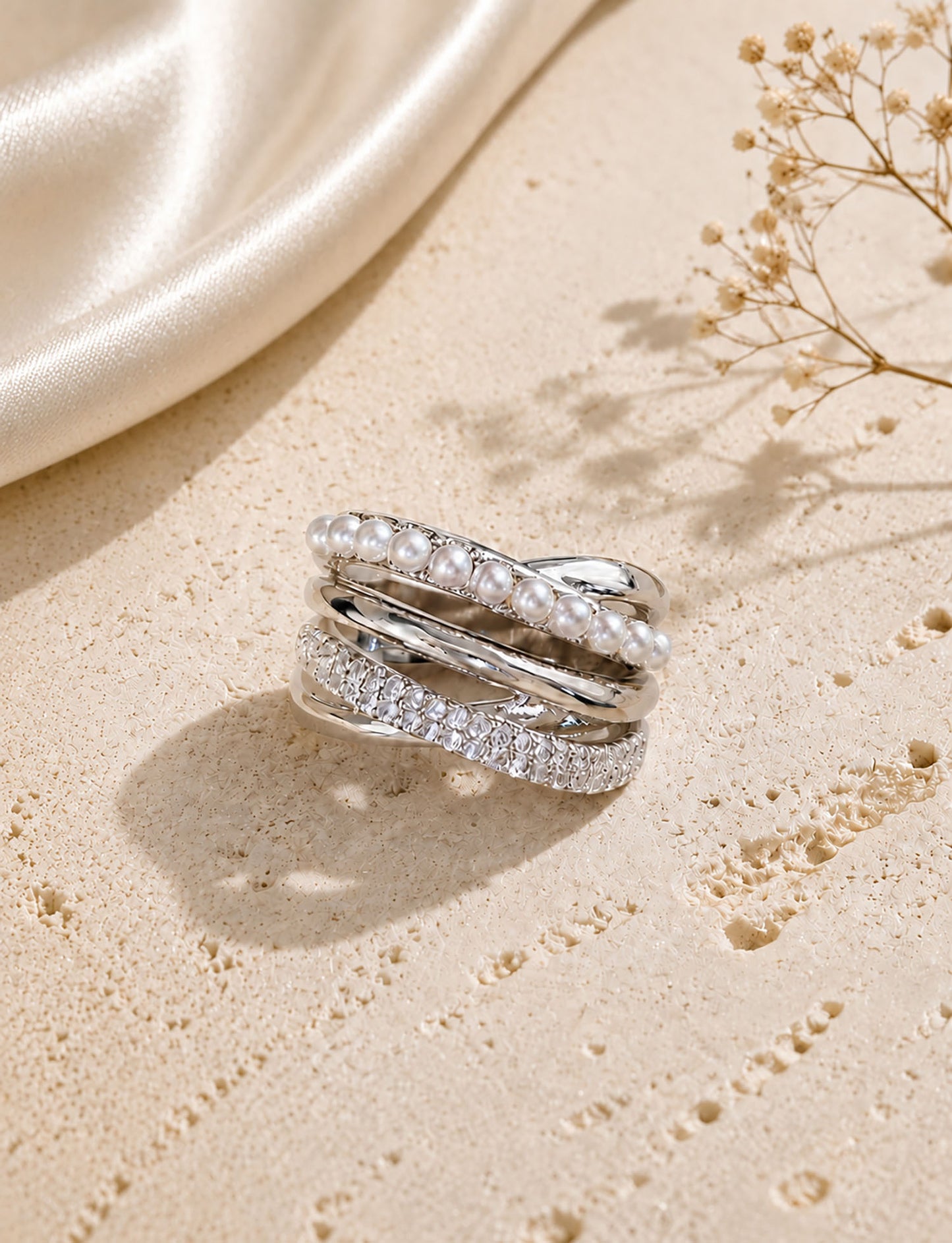 Elegant silver-tone multi-layer crossover ring featuring crystal pearls and micro-paved zirconia resting on a textured aesthetic surface.