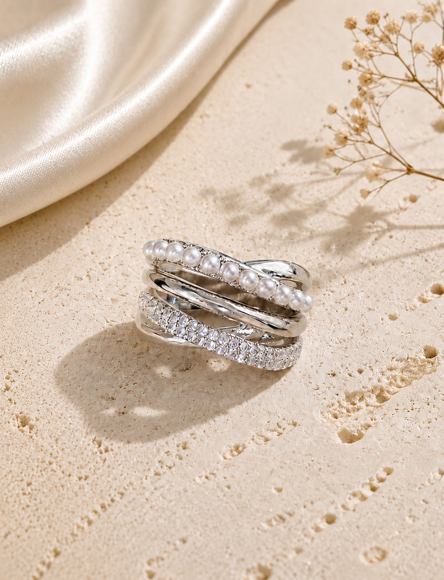 Elegant silver-tone multi-layer crossover ring featuring crystal pearls and micro-paved zirconia resting on a textured aesthetic surface.