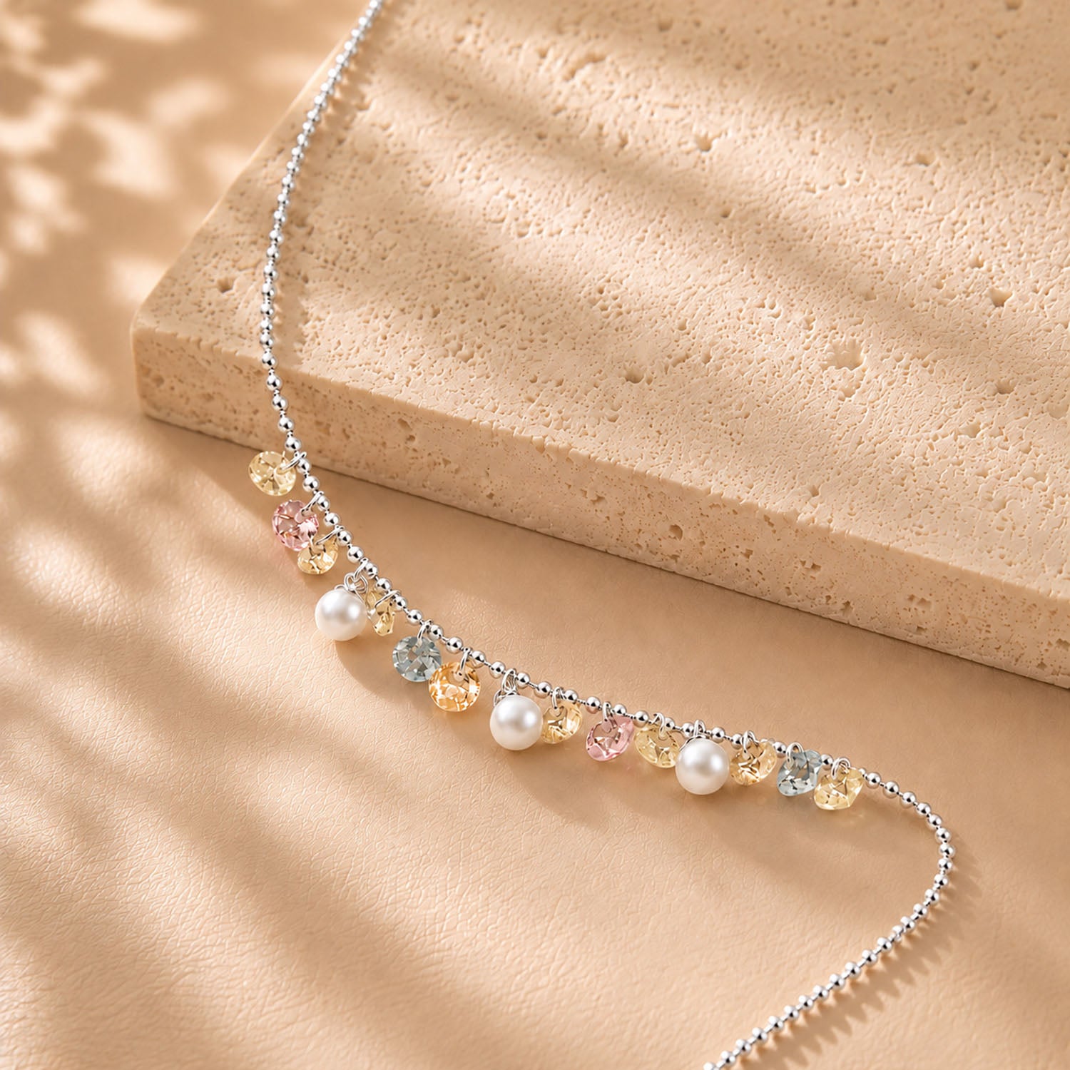 Close up of pastel zirconia and pearl drops on a delicate white gold-tone chain resting on a textured stone surface.
