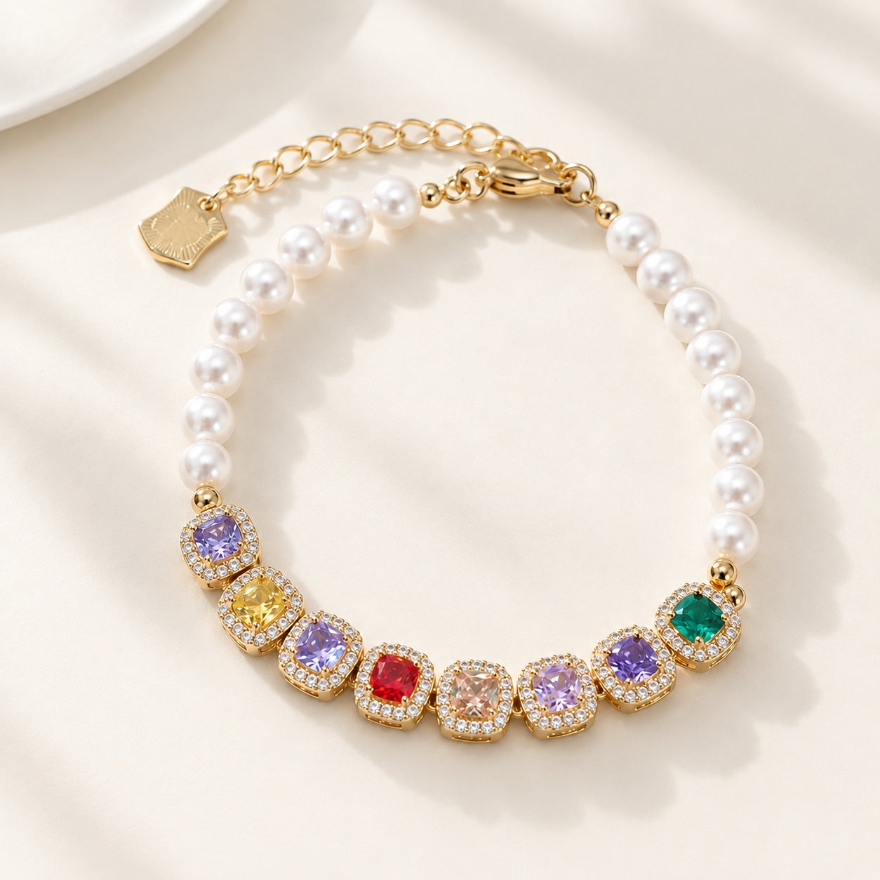 Flatlay view of gold bracelet with colorful cushion cut CZ gems and white pearls on beige background