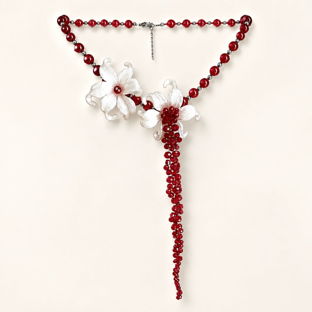 Flat lay of a striking statement necklace featuring deep red beads, white sculpted iris flowers, and a 13cm dramatic beaded tassel on a white background.