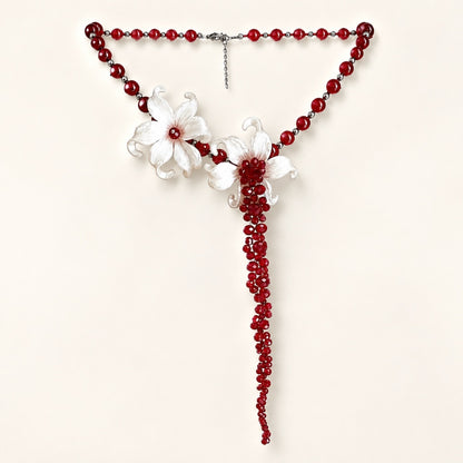 Flat lay of a striking statement necklace featuring deep red beads, white sculpted iris flowers, and a 13cm dramatic beaded tassel on a white background.