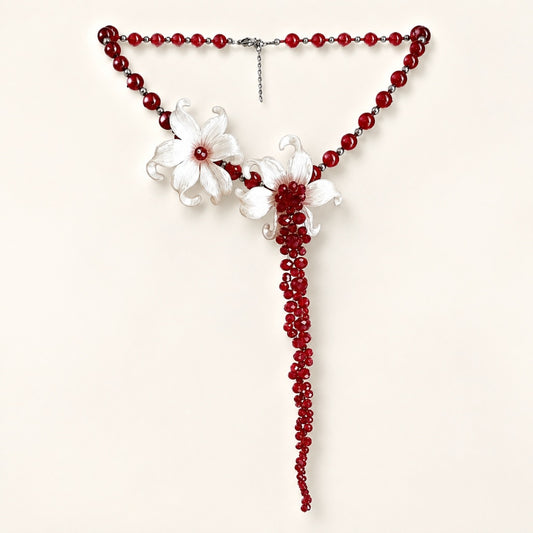 Flat lay of a striking statement necklace featuring deep red beads, white sculpted iris flowers, and a 13cm dramatic beaded tassel on a white background.