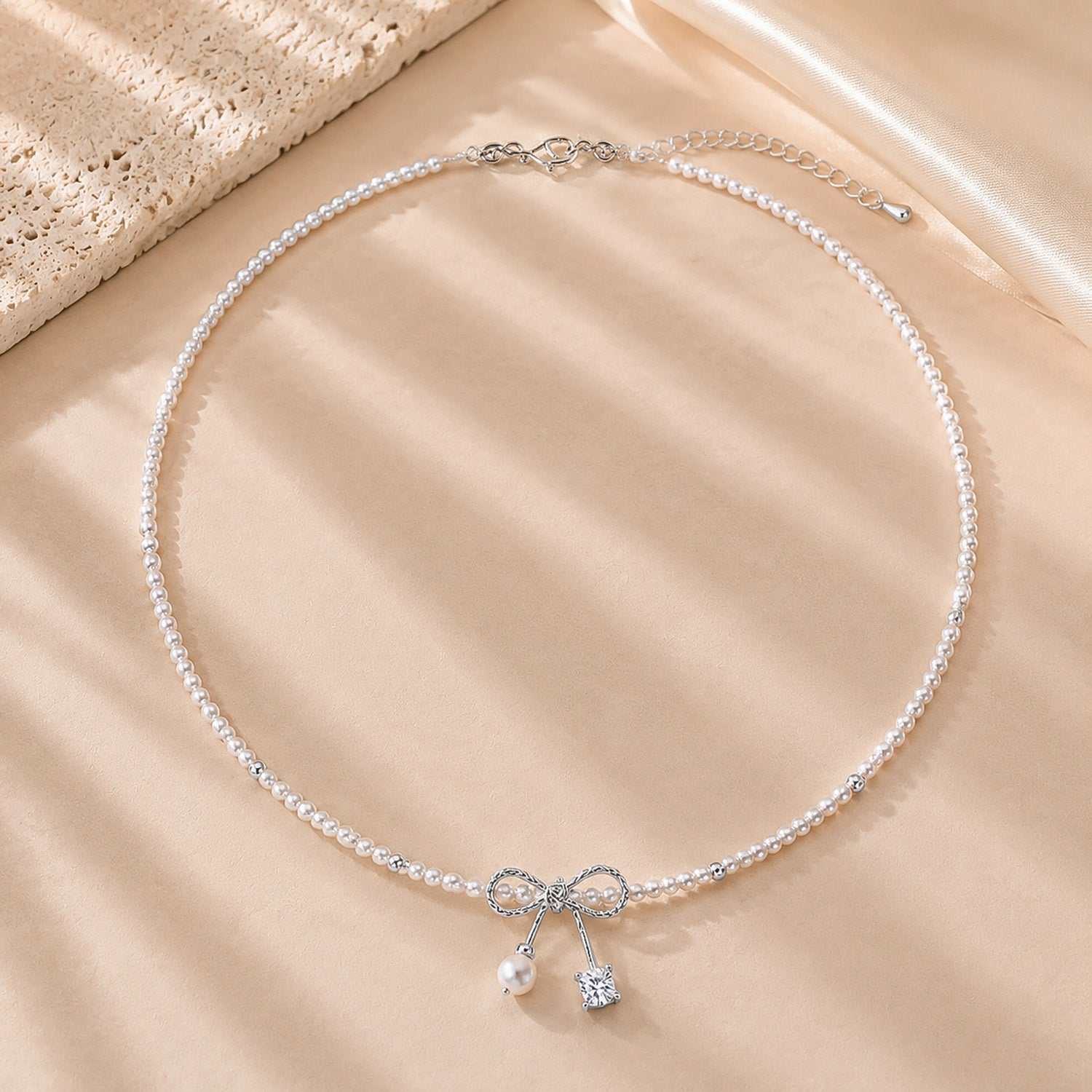Romantic pearl necklace with a white gold-tone ribbon bow resting on textured stone and soft silk.