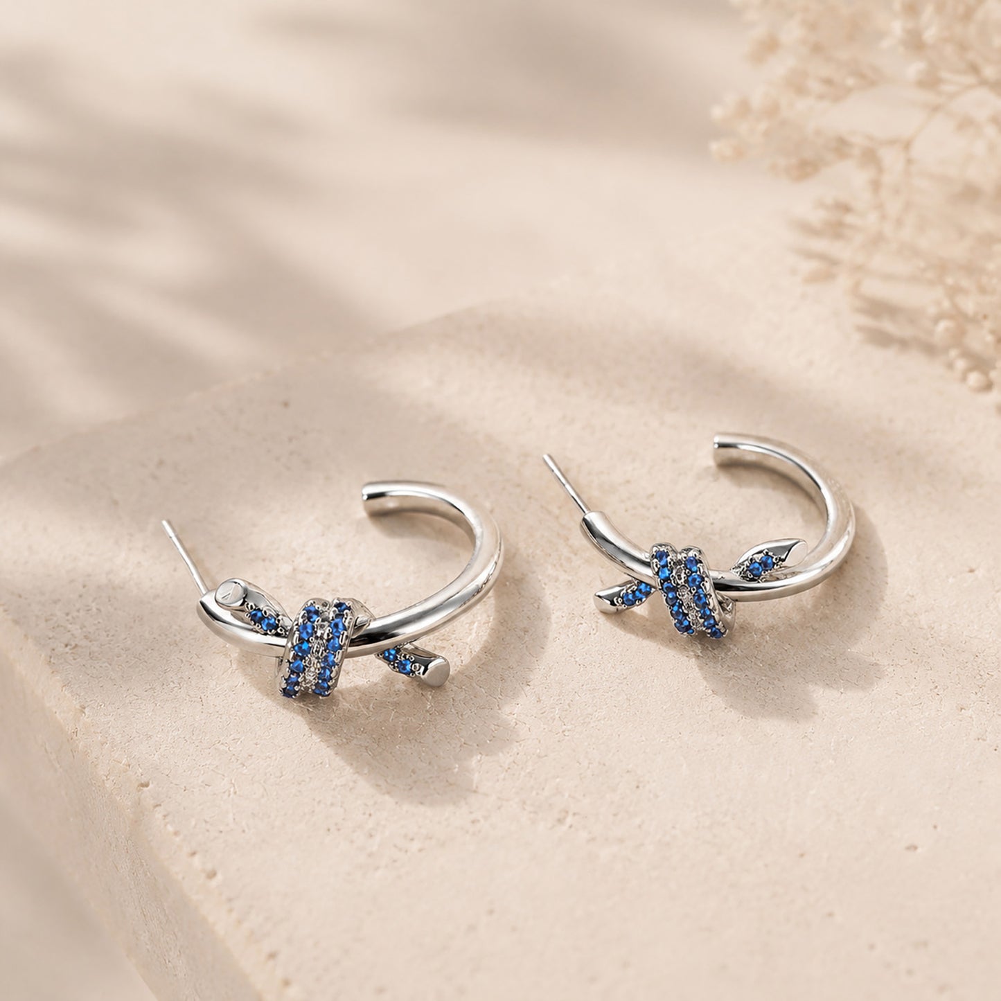Macro close-up of blue zirconia knotted C-hoop earrings resting on a textured stone surface, showcasing 925 silver posts.