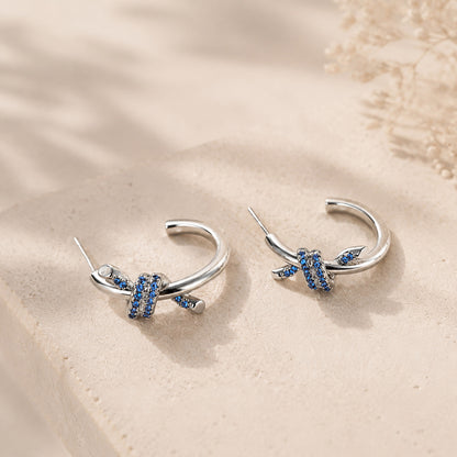 Macro close-up of blue zirconia knotted C-hoop earrings resting on a textured stone surface, showcasing 925 silver posts.