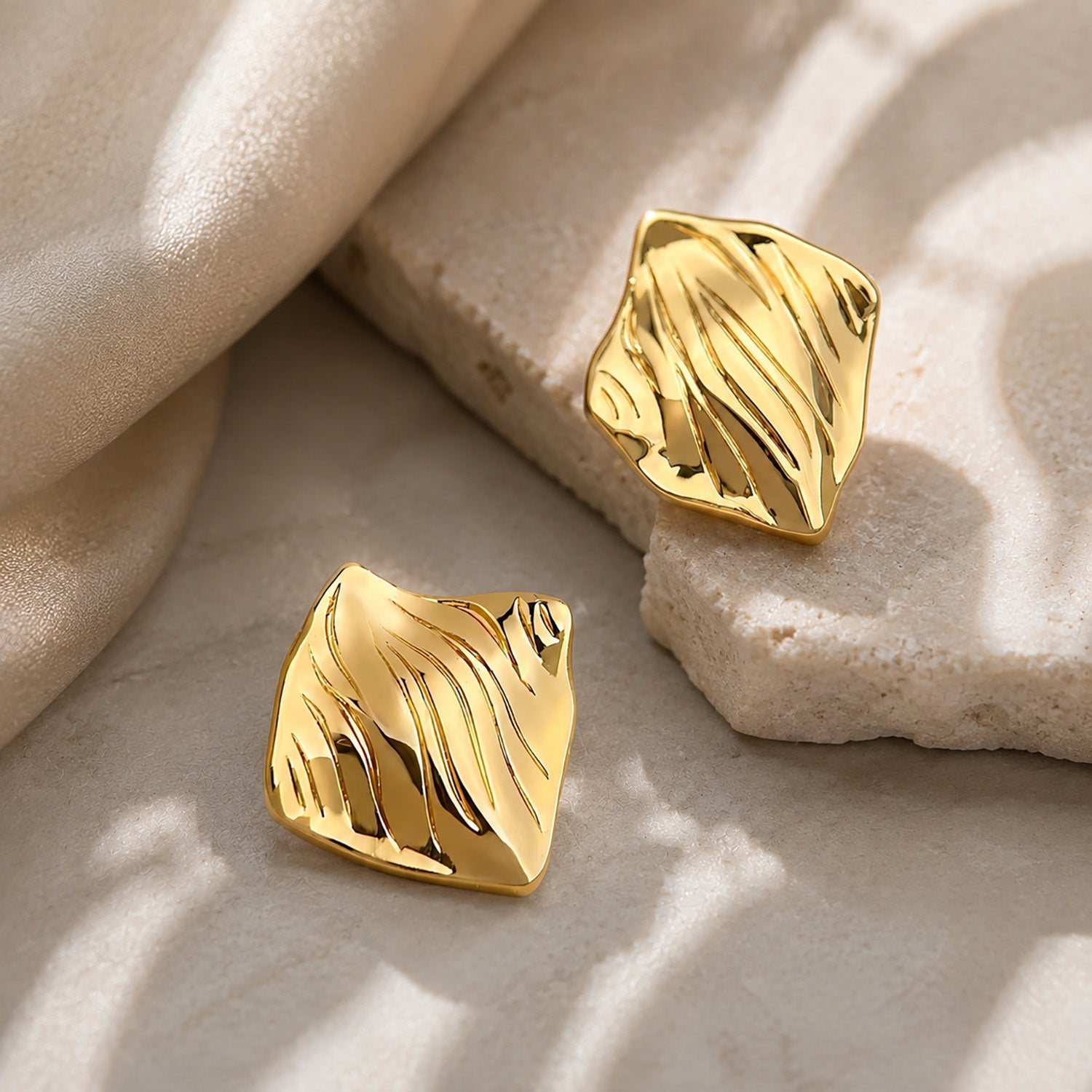 Close-up of bold crinkled square stud earrings showcasing their highly polished, liquid gold-like texture on a textured block.