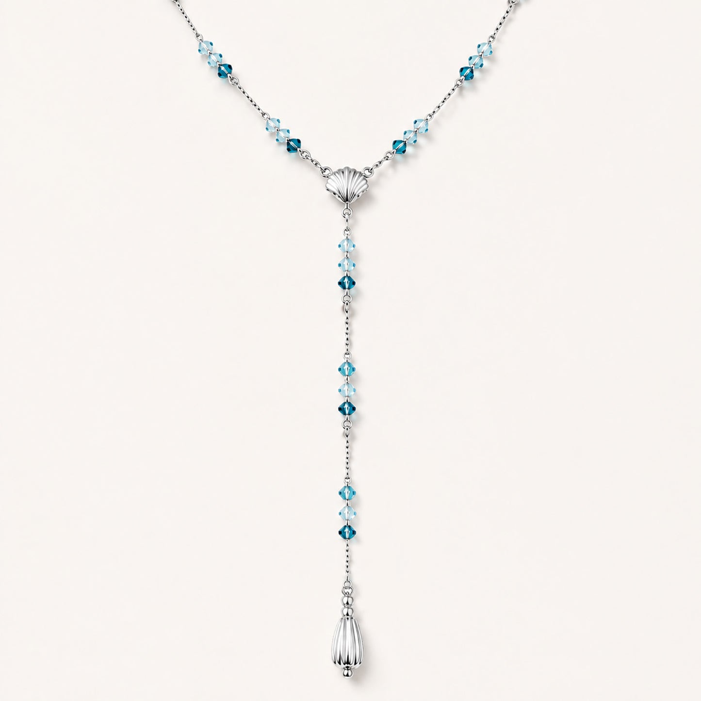 Minimalist flat lay of an elegant Y-necklace featuring a central seashell, blue gemstones, and a teardrop pendant on a white background.