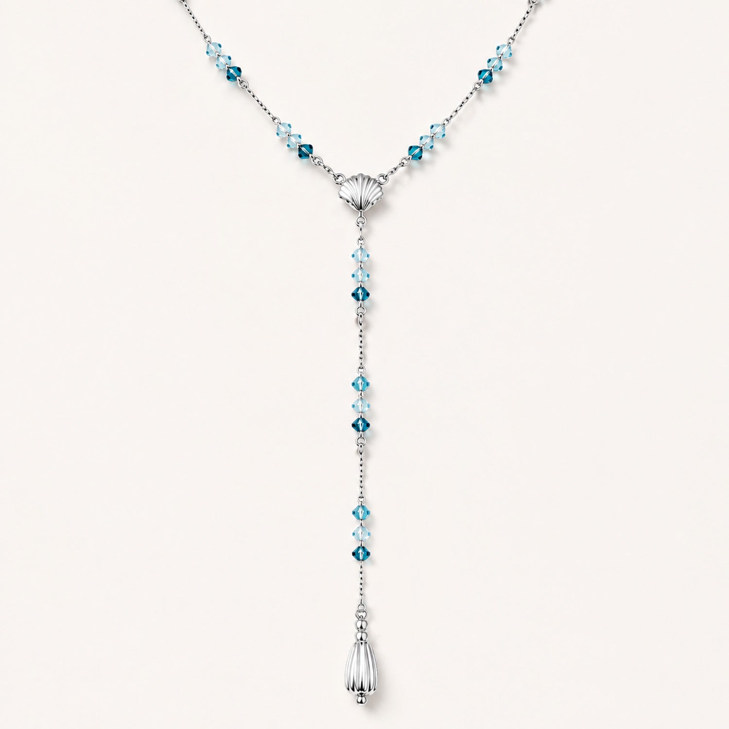 Minimalist flat lay of an elegant Y-necklace featuring a central seashell, blue gemstones, and a teardrop pendant on a white background.