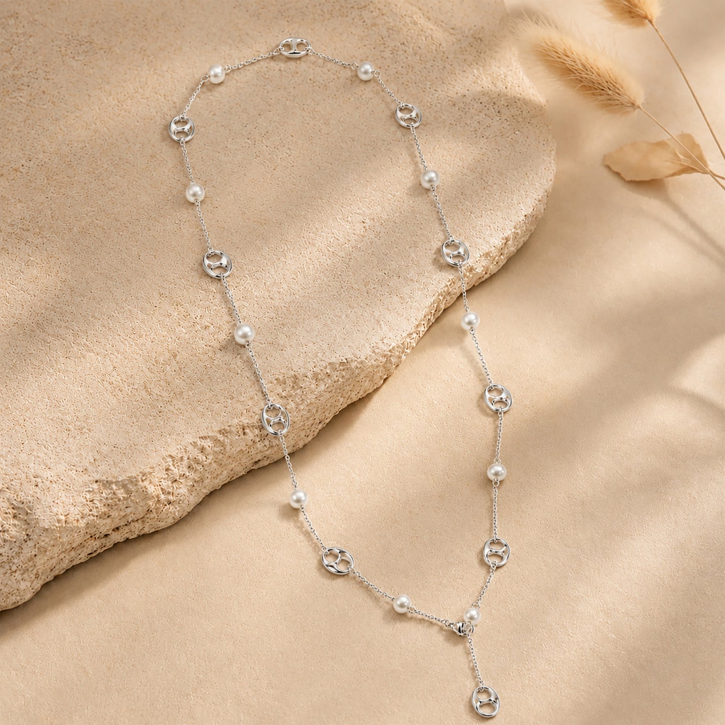 Elegant silver-tone Y-necklace featuring mother-of-pearl and anchor links, resting on a textured stone surface with dried botanicals.