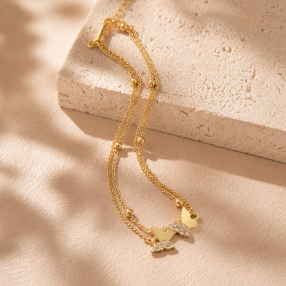 Elegant gold-tone double butterfly anklet draped over a textured stone block in warm sunlight.