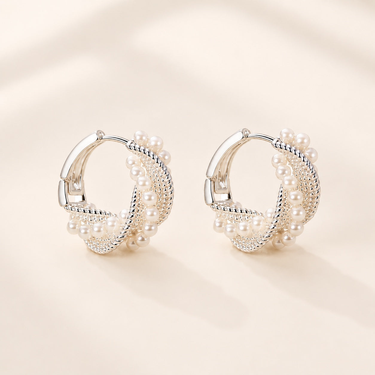 Pair of silver twisted rope pearl huggie hoop earrings displayed on a warm beige background