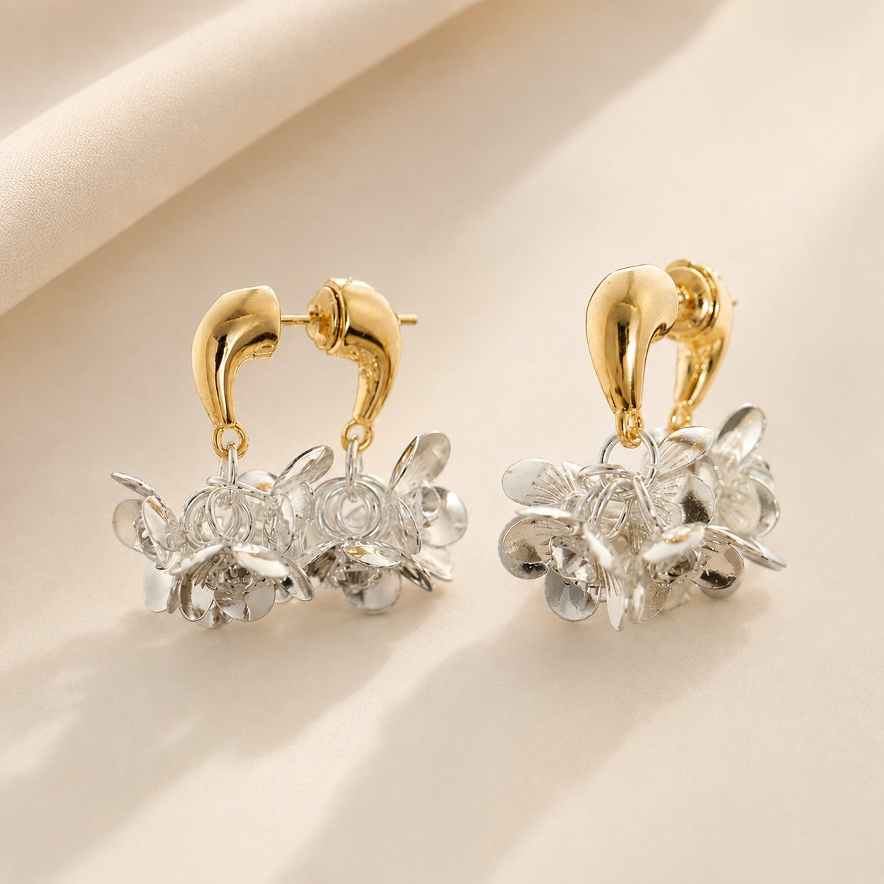 Pair of two-tone gold arch and silver petal floral cluster drop earrings on beige fabric