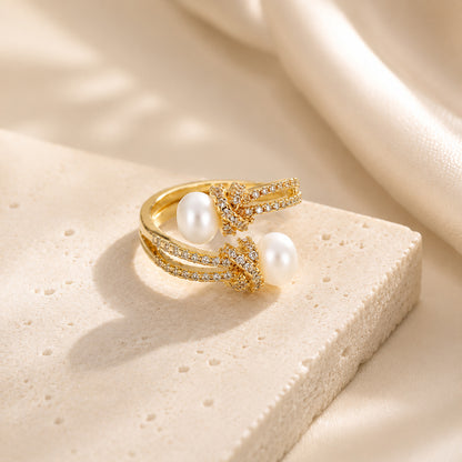 Close-up of an adjustable 18K gold-tone open ring with sparkling zircon knot motifs and freshwater pearls, resting gracefully on textured stone with soft silk.