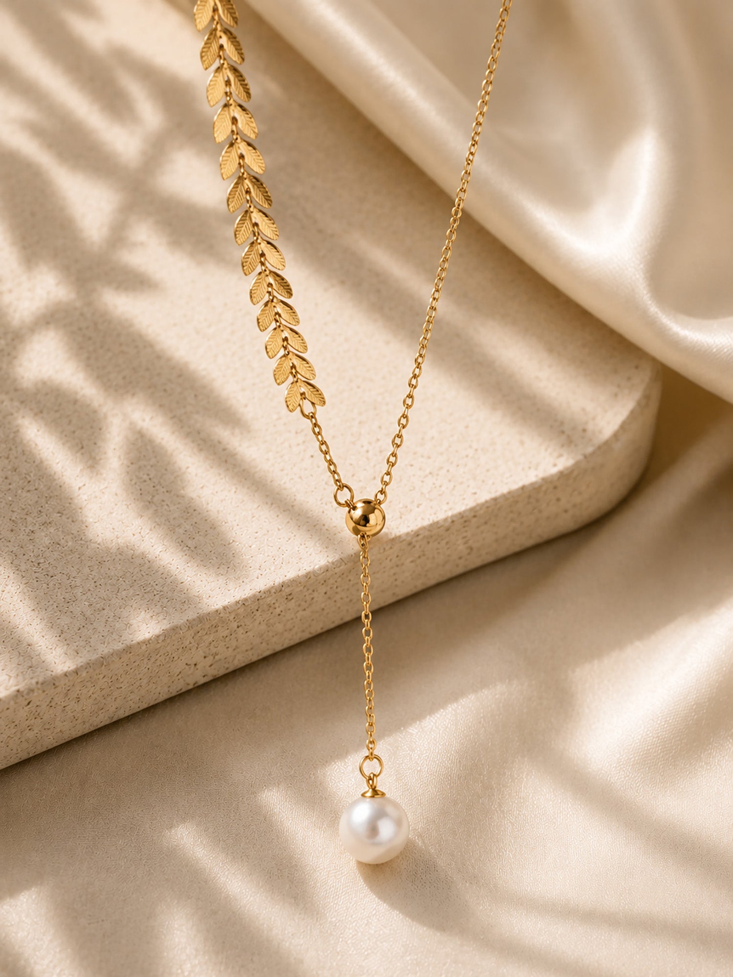 Vintage-inspired gold-tone wheat ear necklace with a pearl drop resting on a textured aesthetic stone surface in soft sunlight.