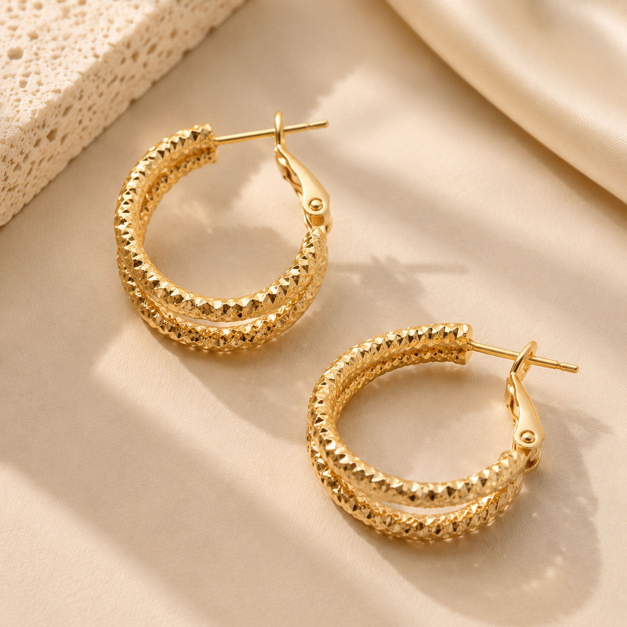 Close up of textured gold-tone hoop earrings resting on warm stone in soft sunlight.