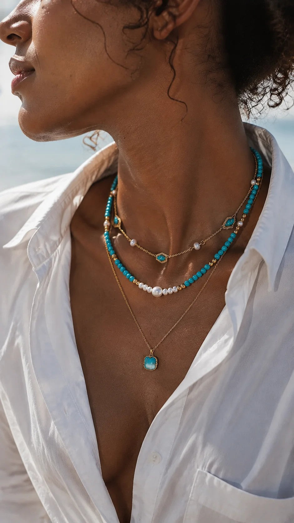 Beautiful woman wearing a crisp white shirt, styling an elegant bohemian layered necklace with turquoise beads and freshwater pearls in warm sunlight.