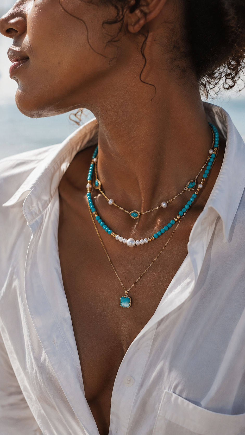 Beautiful woman wearing a crisp white shirt, styling an elegant bohemian layered necklace with turquoise beads and freshwater pearls in warm sunlight.